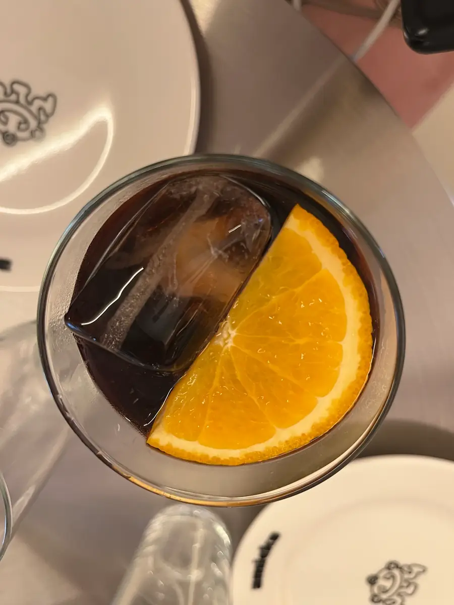 Un cóctel oscuro con hielo y media rodaja de naranja, visto desde arriba